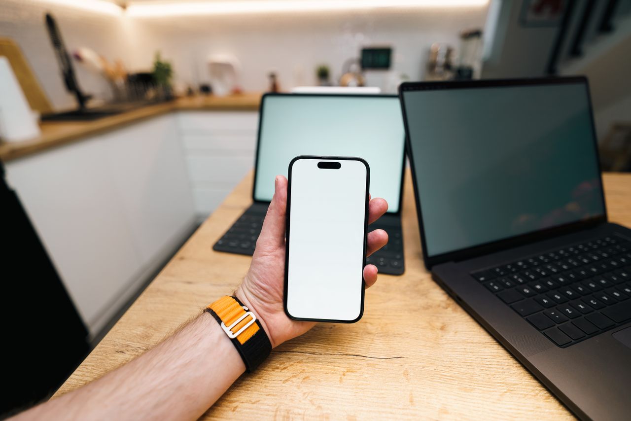 A hand holds a smartphone in a contemporary kitchen with laptops on a wooden table.